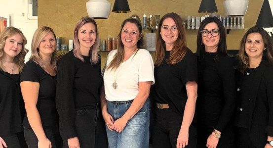 Eine Gruppe von Frauen in schwarzer Kleidung posiert für ein Foto in einem Schönheitssalon mit Regalen voller Produkte im Hintergrund.