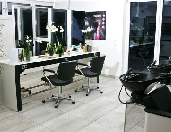 Ein Salon mit einem Schreibtisch und Stühlen vor einer Glaswand. Blumen, Flaschen und andere Gegenstände stehen auf dem Schreibtisch. Rechts befindet sich ein Waschbecken mit Wasserhahn.