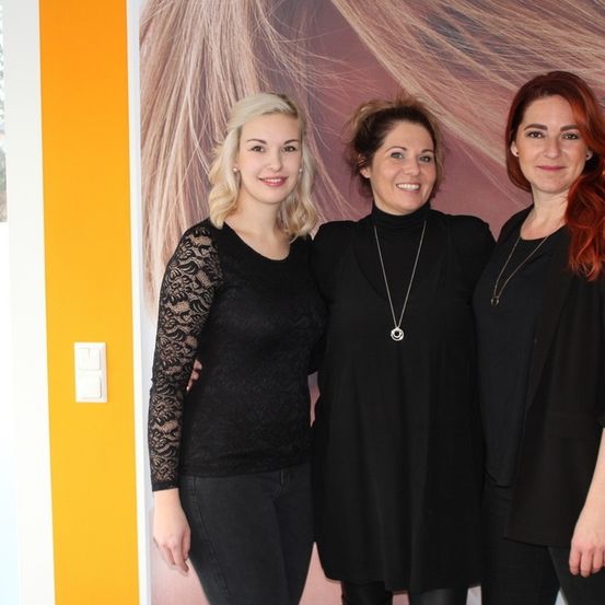 Drei Frauen in schwarzer Kleidung posieren zusammen vor einem Hintergrund mit blondem Haar. Sie tragen alle Halsketten und scheinen zu lächeln. Die Frau auf der linken Seite hat blondes Haar und Ärmel mit Spitze.