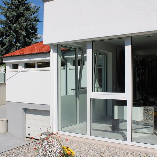 Ein modernes weißes Haus mit einem roten Dach und großen Glastüren und Fenstern. Die Glastüren sind offen und zeigen eine Reflexion eines Baumes und eines Gebäudes.