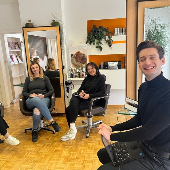 Drei Frauen sitzen in einem Salon, während ein Mann lächelt und mit einem Laptop sitzt. Die Frau auf der linken Seite hält Stift und Papier, und die Frau in der Mitte hält ein Tablet. An der Wand befinden sich Spiegel und Regale mit Büchern und Pflanzen.