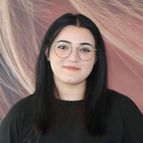 Eine Frau mit langen schwarzen Haaren und runden goldfarbenen Brillengläsern sowie einem schwarzen T-Shirt posiert vor einem rosa Hintergrund.