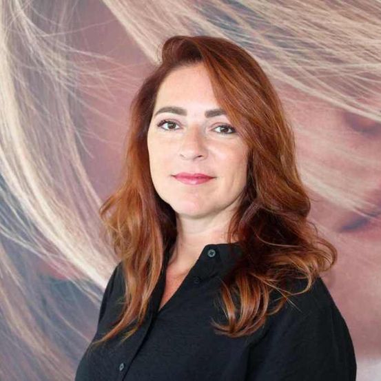 Eine Frau mit roten Haaren lächelt vor einem großen Poster mit dem Haar einer blonden Frau.