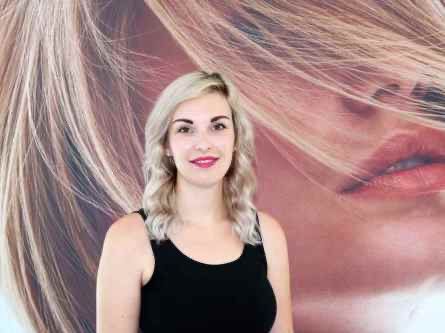 Eine Frau mit blonden Haaren steht vor einem großen Poster einer Frau mit blonden Haaren, beide tragen schwarze trägerlose Oberteile und roten Lippenstift.