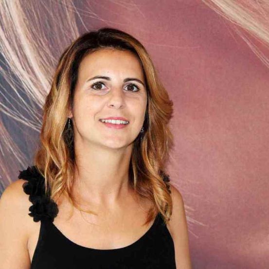 Eine lächelnde Frau mit blonden, gewellten Haaren posiert für ein Foto vor einem rosa Hintergrund. Sie trägt ein schwarzes ärmelloses Top und Ohrringe.