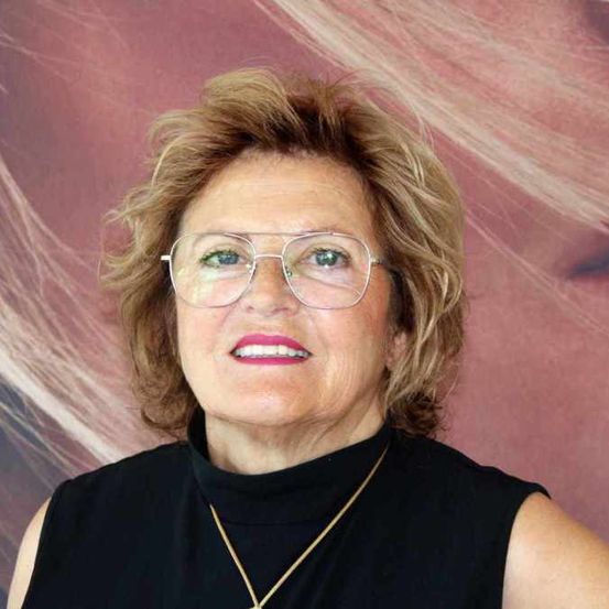 Eine ältere Frau mit kurzen blonden Haaren lächelt in die Kamera. Sie trägt eine Brille, ein schwarzes ärmelloses Oberteil und eine goldene Halskette.