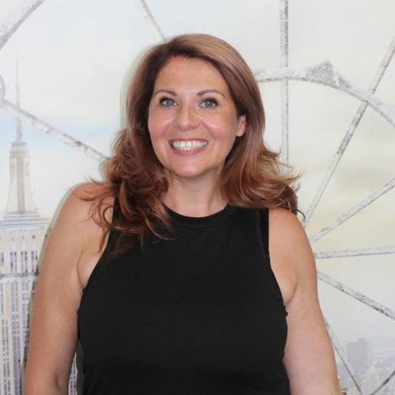 Eine lächelnde Frau mit braunen Haaren in einem schwarzen ärmellosen Oberteil steht vor einem Hintergrund mit einem Rad und dem Empire State Building.