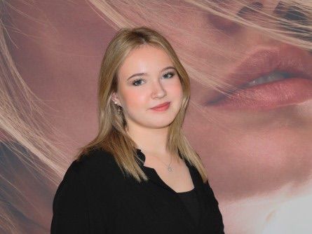 Eine junge Frau mit blonden Haaren steht vor einem Poster. Sie trägt ein schwarzes Oberteil und eine Halskette.