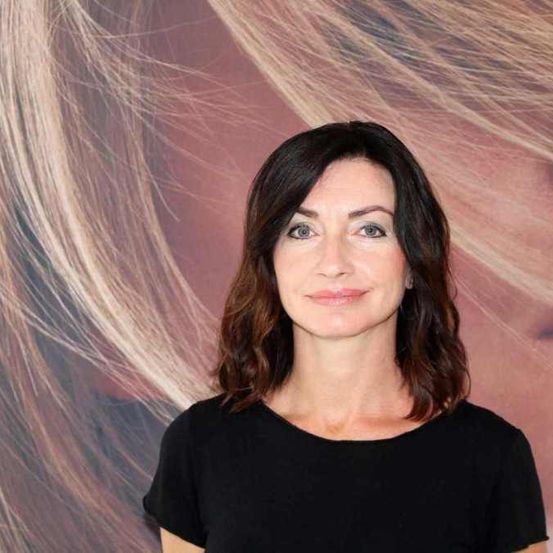 Eine Frau mit dunkelbraunen Haaren und blauen Augen steht vor einem großen Haarposter. Sie trägt ein schwarzes Shirt und lächelt leicht.