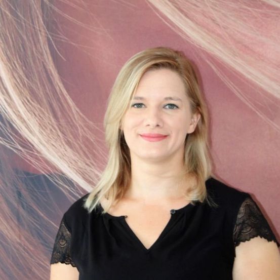 Eine Frau mit blonden Haaren, die ein schwarzes Oberteil trägt, lächelt und posiert für ein Foto vor einem rosa Hintergrund.