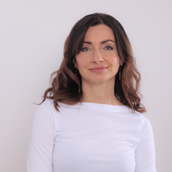 Eine Frau mit braunen, gewellten Haaren, die ein weißes Langarmshirt trägt, lächelt fröhlich in die Kamera.