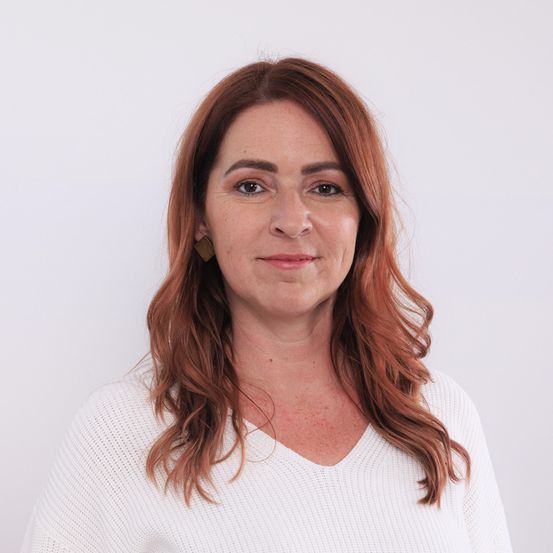 Eine Frau mit braunen, welligen Haaren, die ein weißes Oberteil trägt und in die Kamera lächelt. Sie hat leichtes Make-up und Ohrringe.