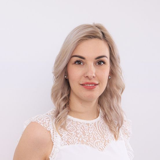 Eine Frau mit blonden Haaren lächelt für ein Foto. Sie trägt ein weißes Spitzenoberteil. Der Hintergrund ist weiß.