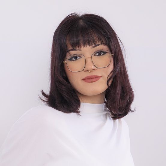Eine Frau mit braunem Haar, Pony und Brille posiert für ein Foto. Sie trägt ein weißes Rollkragenshirt. Ihr Make-up umfasst Eyeliner, Lidschatten und rosa Lippenstift.