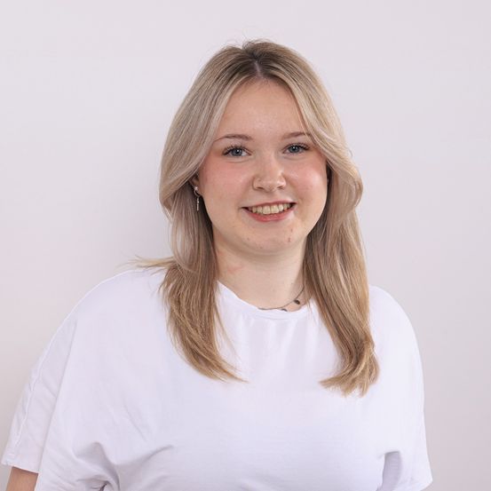 Eine Frau mit blonden Haaren und einem weißen T-Shirt lächelt in die Kamera. Sie trägt Ohrringe und eine Halskette.