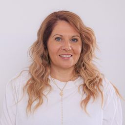 Eine Frau mit blonden welligen Haaren trägt ein weißes Shirt. Sie hat eine Halskette mit Sonnenanhänger. Sie lächelt und scheint für ein Foto zu posieren.