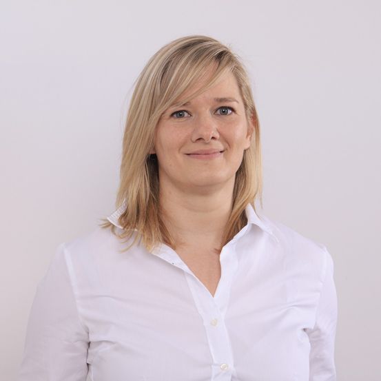 Eine Frau mit blonden Haaren lächelt und posiert für ein Foto. Sie trägt ein weißes Hemd.