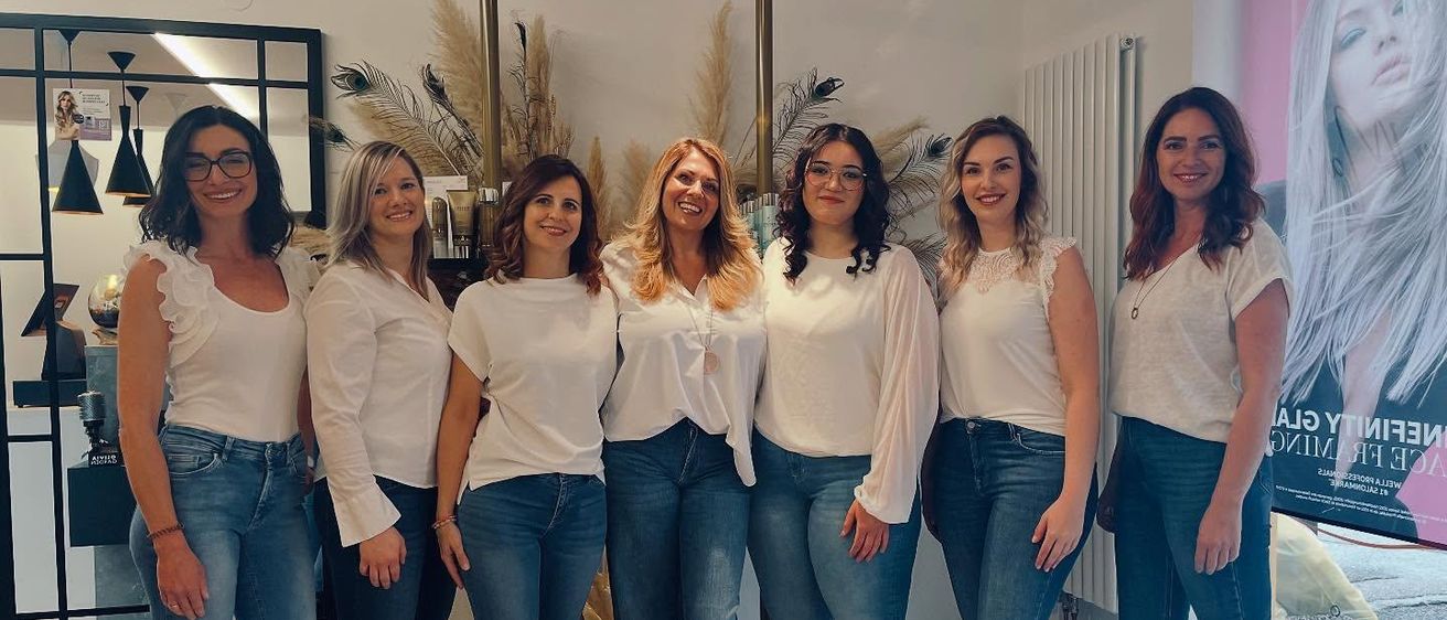 Sieben Frauen posieren für ein Foto in einem Raum. Sie tragen alle weiße Blusen und blaue Jeans. Sie lächeln und scheinen glücklich zu sein.