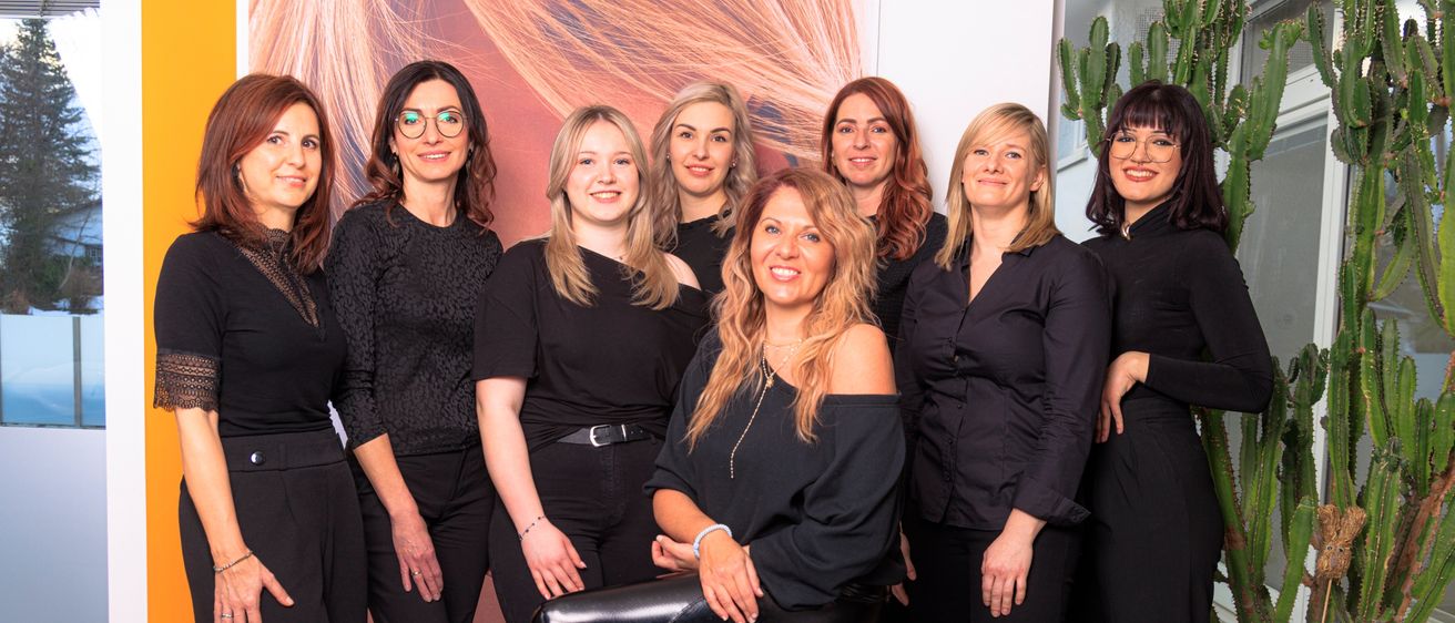 Eine Gruppe von Frauen in schwarzen Outfits steht in einem Studio zusammen. Die Frau in der Mitte sitzt auf einem Stuhl und lächelt. Ein großes Haarbild steht hinter ihnen.