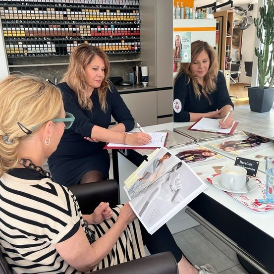 Drei Frauen sitzen in einem Salon, zwei schreiben auf Papiere und eine liest. Eine Kaffeetasse und Zeitschriften sind auf dem Schreibtisch. Dahinter steht ein Regal mit vielen Flaschen.