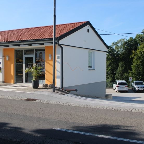 Ein kleines weißes Gebäude mit rotem Dach steht an einer Straße. Zwei Autos sind davor geparkt. Vor dem Gebäude stehen zwei Topfpflanzen.