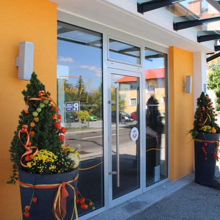 Der Eingang zu einem Gebäude hat Glastüren mit dekorativen Pflanzen und Bändern. Das Gebäude hat gelbe Wände und ein Schild mit dem Namen 'Frausens'. Es gibt auch zwei Wandlampen.