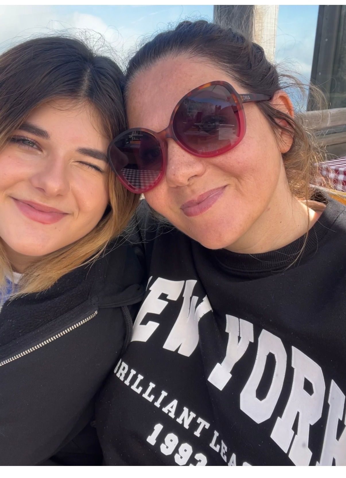 Two women smiling and posing for a selfie. The woman on the left has blonde hair, and the woman on the right is wearing sunglasses. They both wear black jackets.