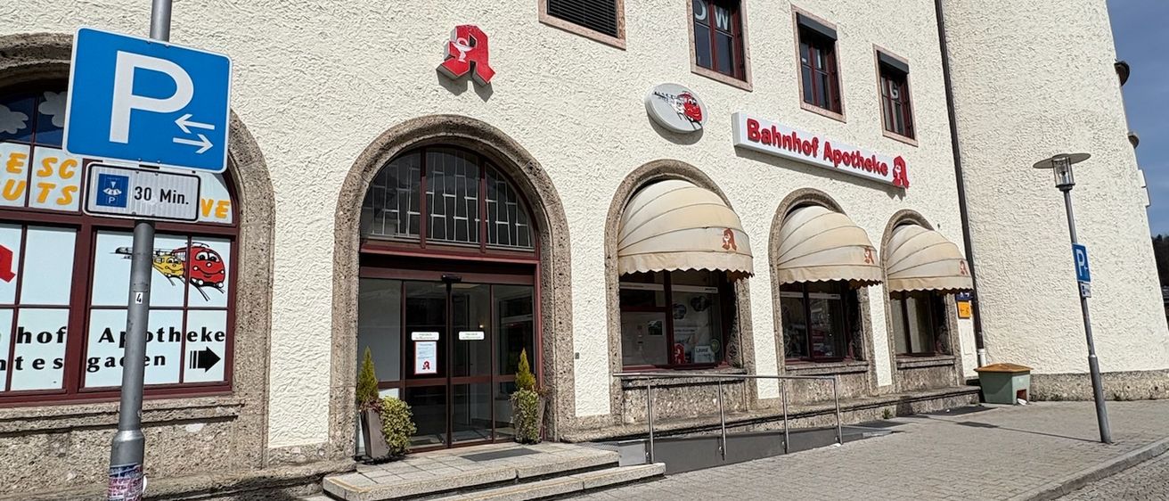 Ein großes Gebäude mit Bogeneingängen, von denen einer ein Schild mit der Aufschrift 'Bahnhof Apotheke' hat. In der Nähe befindet sich ein Parkzeichen.