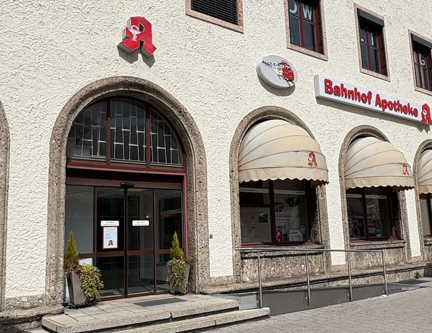 Ein großes Gebäude mit Bogeneingängen, von denen einer ein Schild mit der Aufschrift 'Bahnhof Apotheke' hat. In der Nähe befindet sich ein Parkzeichen.