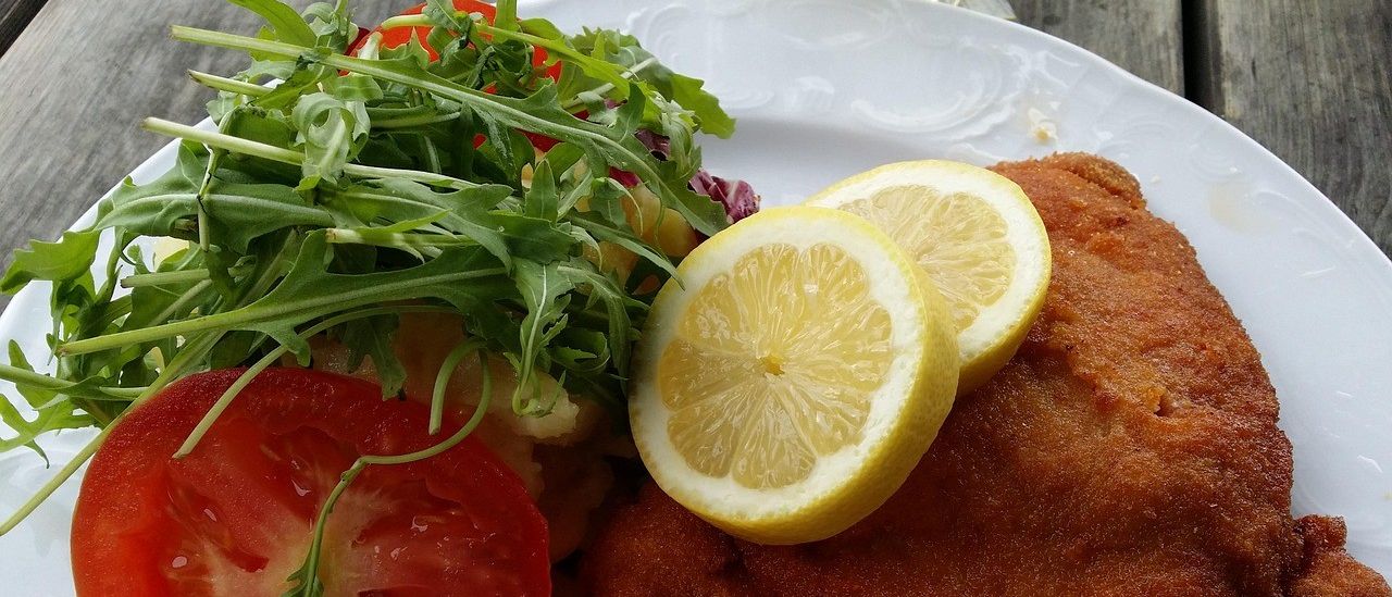 Ein Teller mit paniertem Hähnchen, Zitronenscheiben, Salat und einer kleinen Schüssel Marmelade.