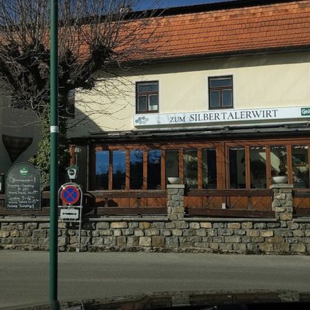 Ein Gebäude mit einem roten Dach und einem Schild, das 'Zum Silbertalerwirt' lautet. Das Gebäude hat große Fenster und einen Holzzaun. Es gibt eine Tafel mit Schrift darauf.