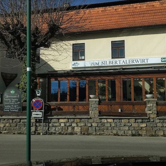 Ein Gebäude mit einem roten Dach und einem Schild, das 'Zum Silbertalerwirt' lautet. Das Gebäude hat große Fenster und einen Holzzaun. Es gibt eine Tafel mit Schrift darauf.
