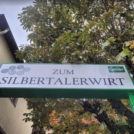 Ein Straßenschild mit der Aufschrift 'Zum Silbertalerwirt' ist an einem Baum befestigt. Es hat einen grünen Rand und ein Logo von Gösser.