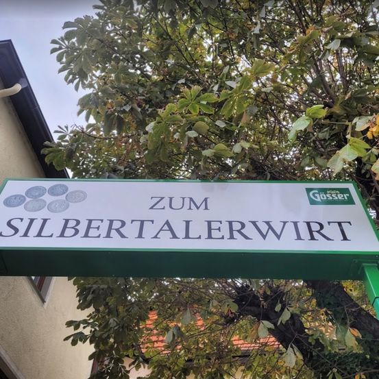 Ein Straßenschild mit der Aufschrift 'Zum Silbertalerwirt' ist an einem Baum befestigt. Es hat einen grünen Rand und ein Logo von Gösser.