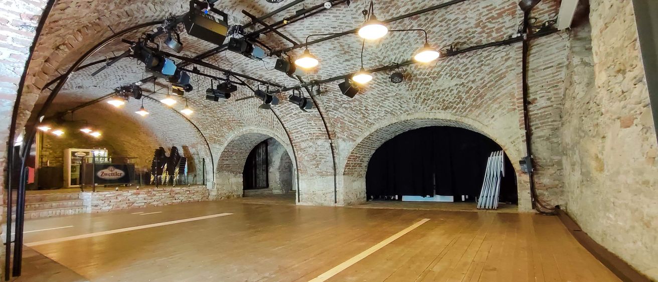 Ein leeres Theater mit Holzboden und Ziegelwänden, mehrere Lampen an der Decke und zwei gewölbte Türen, die zu anderen Räumen führen.