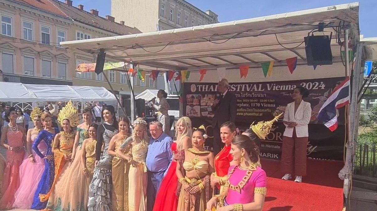 Eine Gruppe von Menschen, darunter Frauen in traditionellen Kostümen und ein Mann, stehen auf einem roten Teppich, wahrscheinlich bei einem Festival. Hinter ihnen befindet sich eine Bühne mit einem Zelt, Flaggen und Bannern. Eine Frau singt in ein Mikrofon.