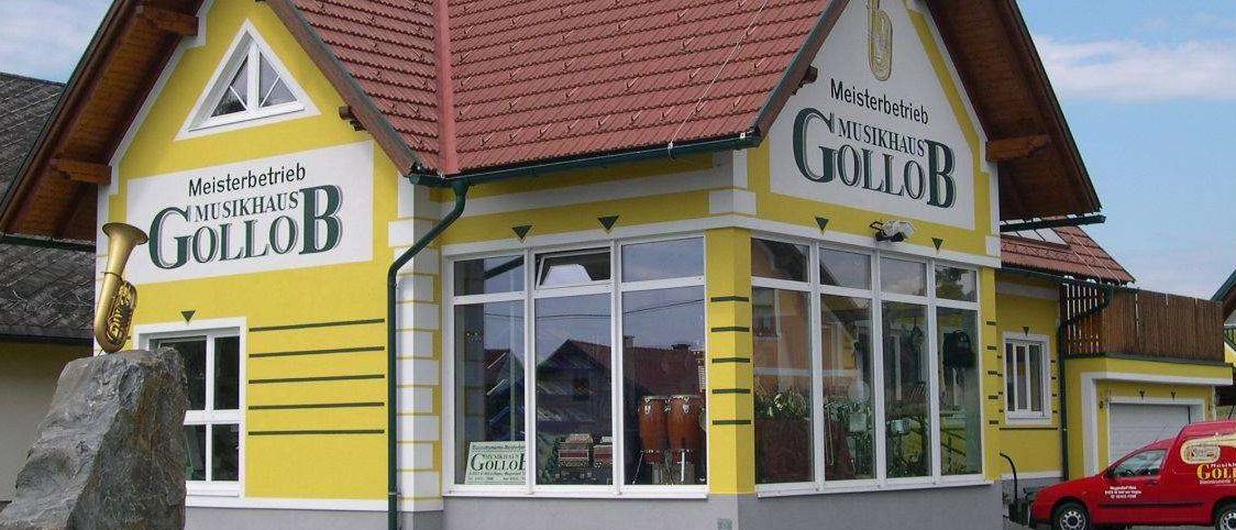 Ein gelbes Gebäude mit rotem Dach namens 'Gollob'. Es hat große Fenster, einen Stein davor und ein rotes Auto, das in der Nähe geparkt ist.