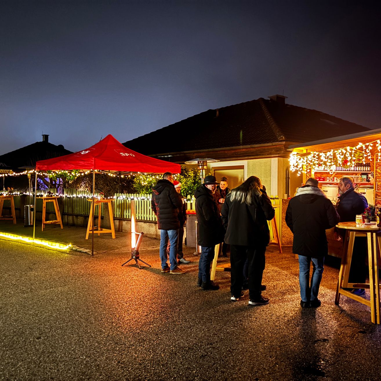 Mehrere Personen versammeln sich am Abend bei einer Outdoor-Veranstaltung, unter einem roten Zelt und verschiedenen Lichtern. Einige stehen vor einer Bartheke mit Getränken.