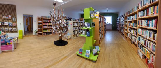 Innenansicht einer Bibliothek mit Holzböden und Bücherregalen. Eine Baum mit Büchern in der Mitte. Ein grünes Regal ist rechts.