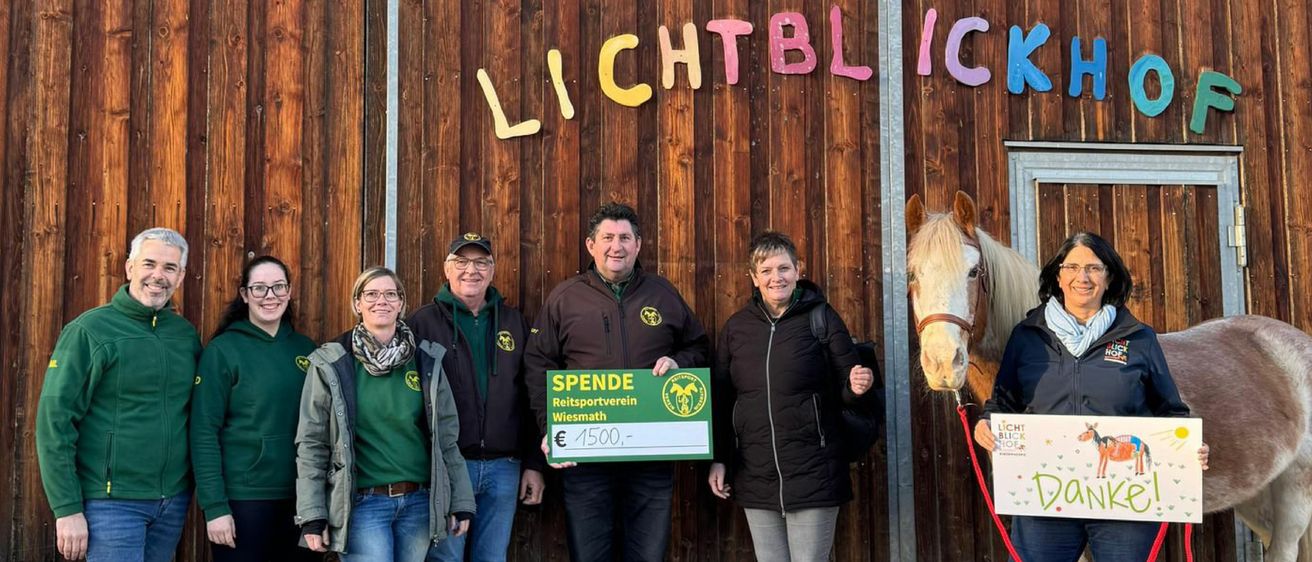 Vier Personen stehen vor einer holzverkleideten Wand mit der Aufschrift 'Lichtblick', ein Mann hält einen grünen Scheck über 1500 Euro. Die Gruppe lächelt und ein Pferd steht in der Nähe.