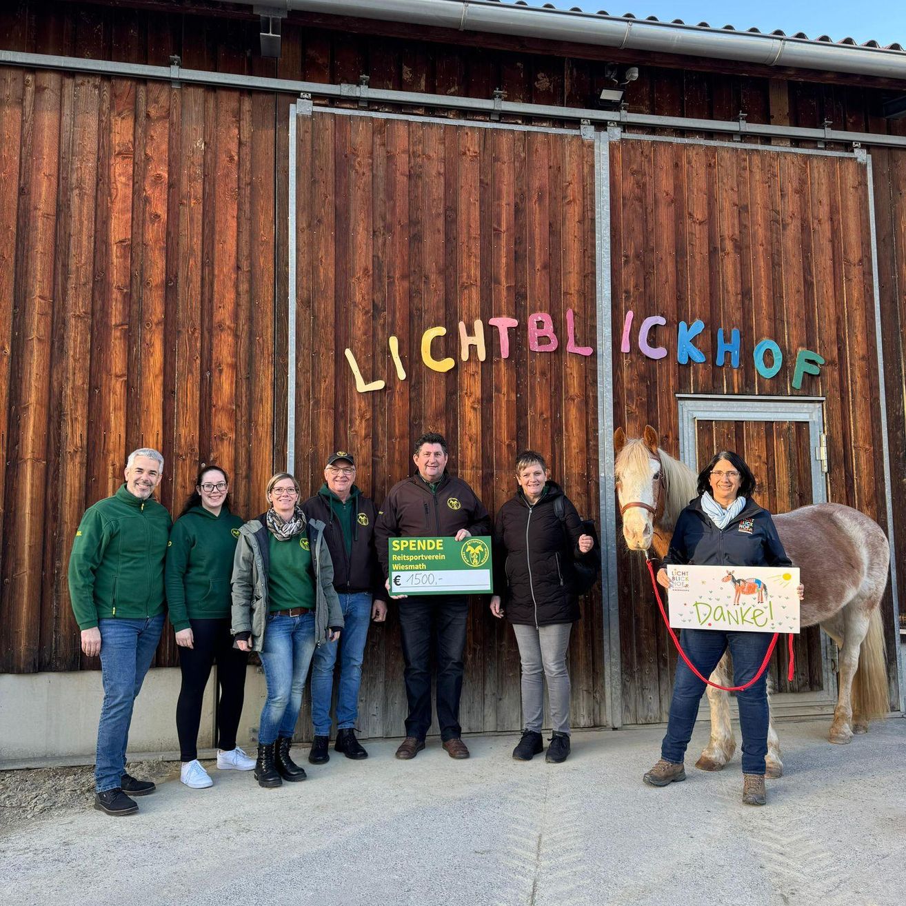 Eine Gruppe von Menschen steht vor einer Scheune mit einem Pferd. Sie halten einen grünen Scheck und ein Schild mit der Aufschrift Danke! Die Scheune hat eine Holzwand und eine Tür.