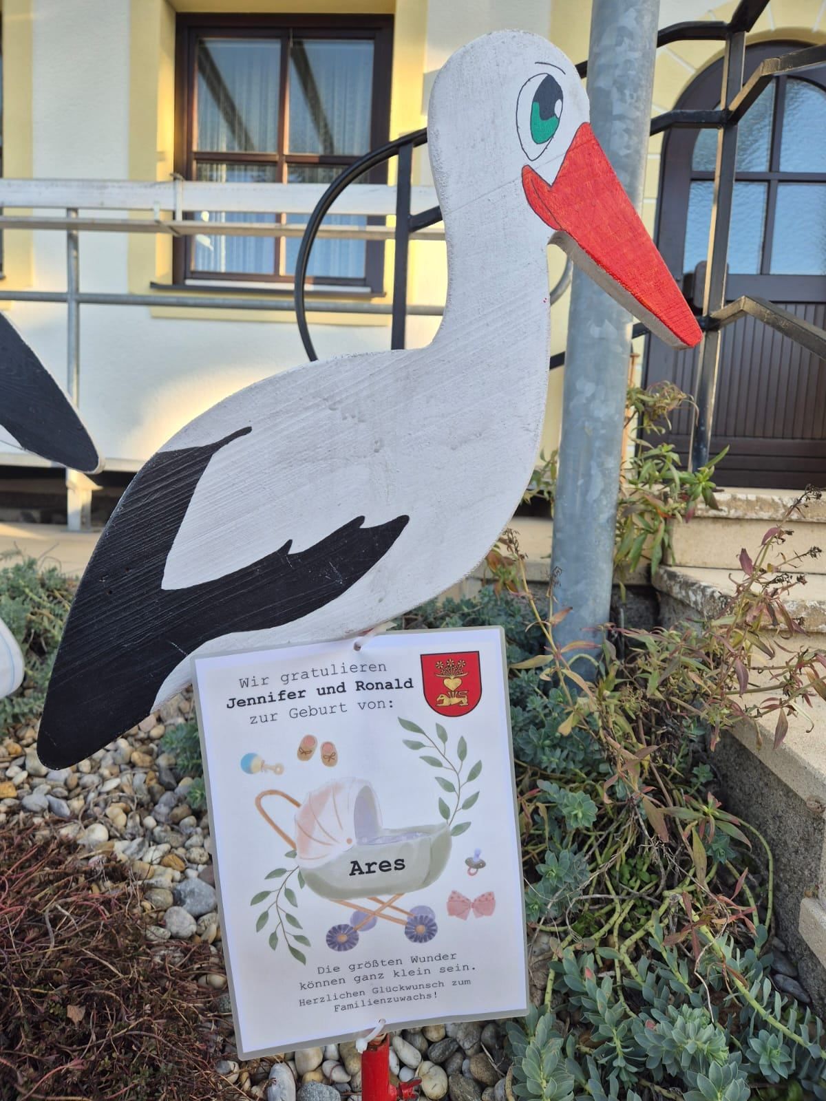 Eine Storchstatue steht vor einem Haus, in der Nähe einer Treppe mit einer Pflanze. Ein Karton mit einem Kinderwagen und dem Namen Ares ist davor platziert.