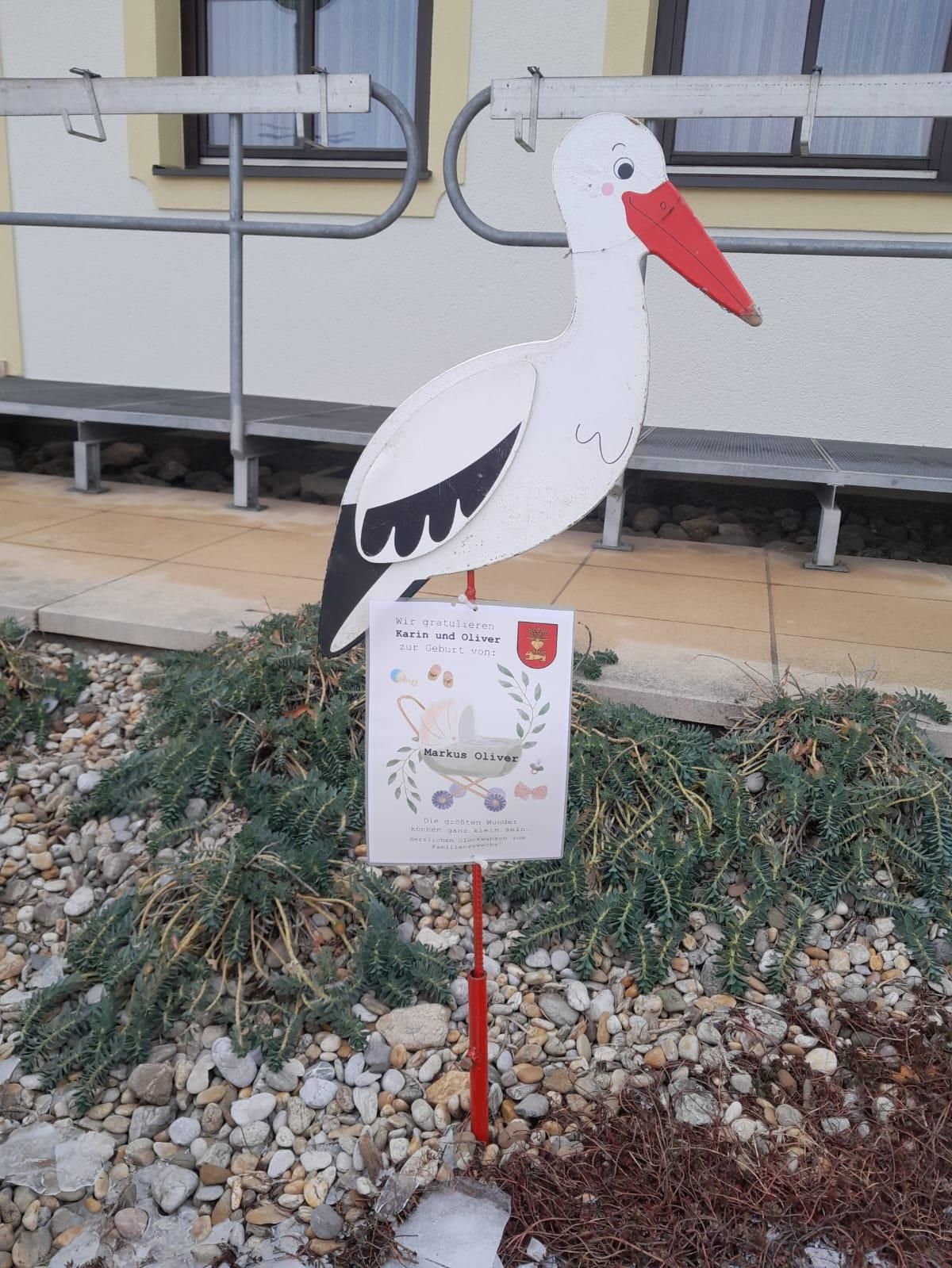 Eine Storchstatue steht in einem Gartenbereich mit einer Karte, auf der steht 'Wir gratulieren Karin und Oliver'. Hinter der Statue befindet sich ein Gebäude mit einer Bank und einer Treppe.