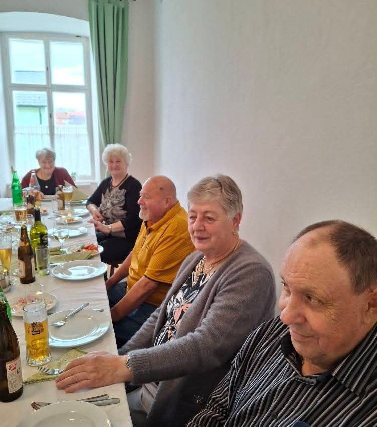 Gruppe älterer Menschen sitzt zu einer Mahlzeit an einem Tisch. Der Tisch ist mit Tellern, Gläsern und Flaschen gedeckt. Sie lächeln und schauen zur Kamera. Dahinter ist ein Fenster mit grünen Vorhängen zu sehen.