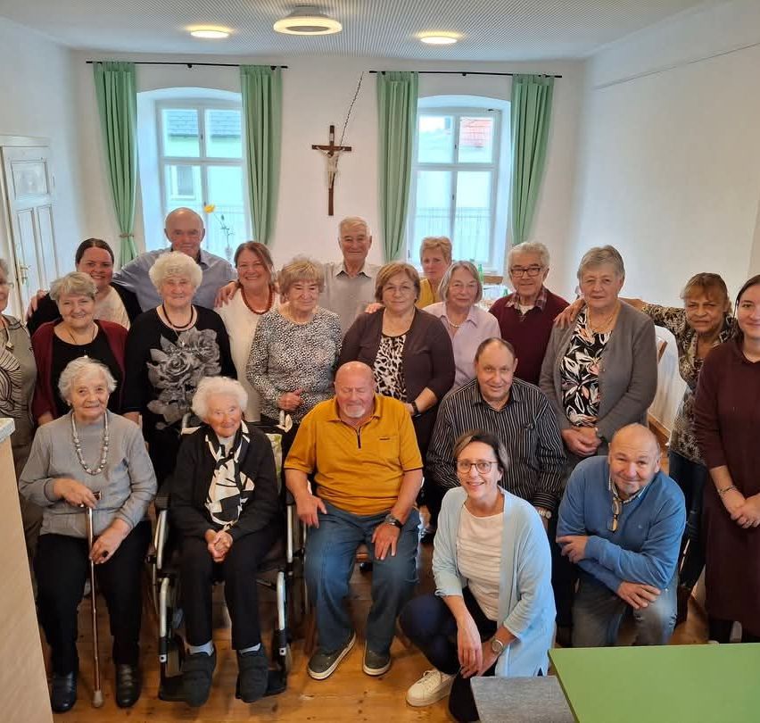 Eine Gruppe älterer Menschen, einige in Rollstühlen, posiert für ein Foto in einem Raum mit Holzböden, weißen Wänden und grünen Vorhängen. Ein Kreuz hängt an der Wand.