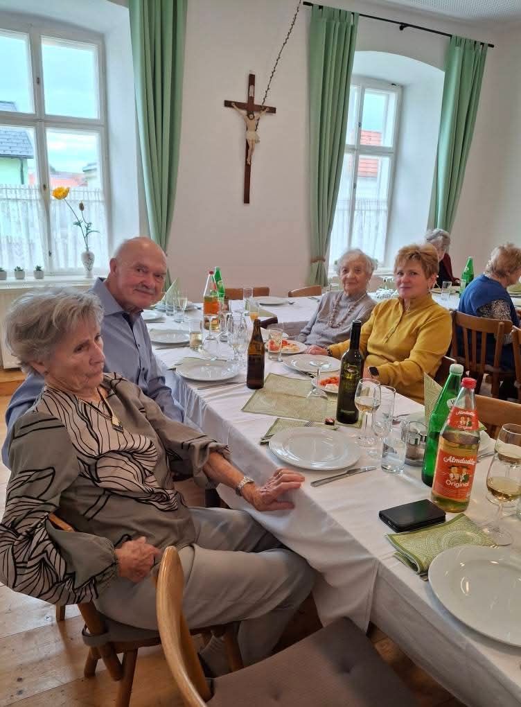 Eine Gruppe älterer Menschen versammelt sich um einen Esstisch und lächelt. Ein Kreuz ist an der Wand hinter ihnen angebracht. Flaschen und Gläser stehen auf dem Tisch. Der Raum hat weiße Wände und grüne Vorhänge.