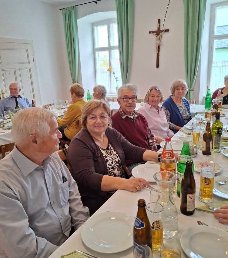 Eine Gruppe älterer Personen sitzt lächelnd um einen Esstisch herum. Flaschen, Gläser und Teller sind auf dem Tisch. Ein Kreuz befindet sich an der Wand. Ein Blumenstrauß in einer Vase steht in der Nähe des Fensters.