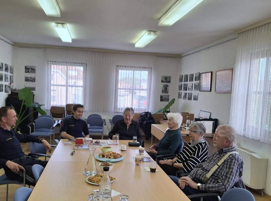 Eine Gruppe von Menschen in einem Besprechungsraum, einige sitzen um einen Tisch mit Essen, andere stehen. Zwei Frauen mit weißen Haaren sitzen in der Nähe und lächeln. Helle Lampen hängen an der Decke.