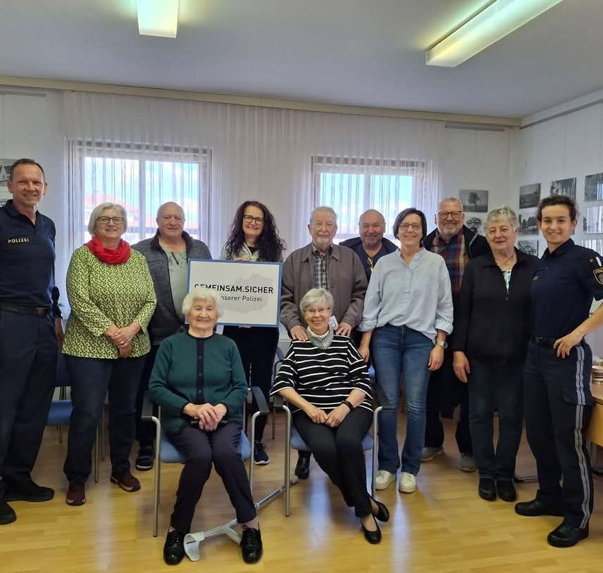 Eine Gruppe von Menschen steht und sitzt in einem Raum, in dem zwei Frauen eine Auszeichnung erhalten. Sie alle lächeln und posieren für ein Foto. Die Frau in der Mitte hält ein Zertifikat.