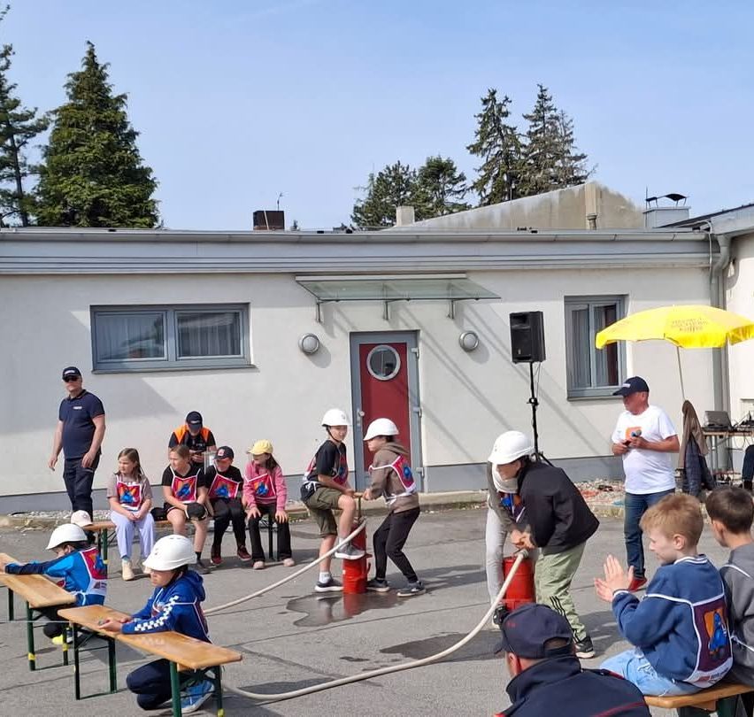 Eine Gruppe von Kindern und Erwachsenen versammelt sich vor einem Gebäude. Sie tragen Helme und halten eine Schläuche. Ein Mann sprüht Wasser aus einem Hydranten.